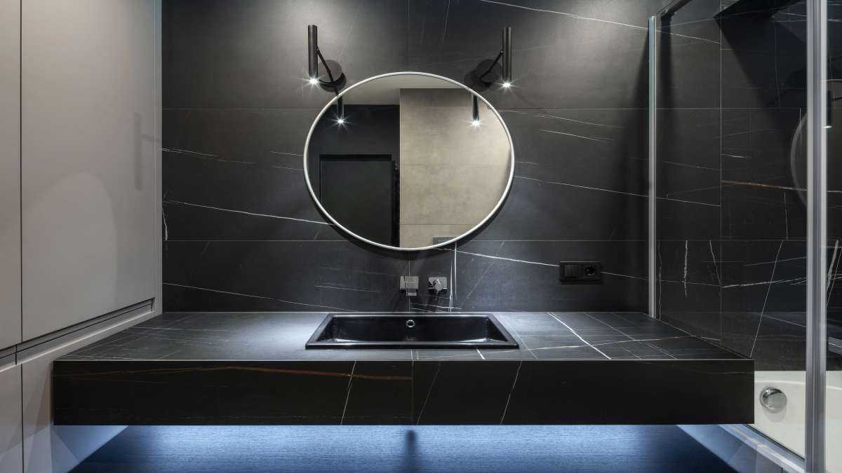 Een zwarte inbouw wastafel met overloopgat onder een ronde spiegel op een zwart tafelblad in een moderne badkamer.