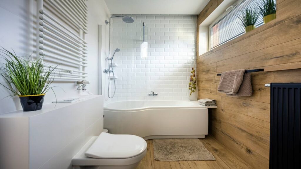 Een kleine badkamer met een witte bad en douche combinatie links van een houten wand die doorloopt op de grond waarop nog voor de douche een wit toilet staat. 