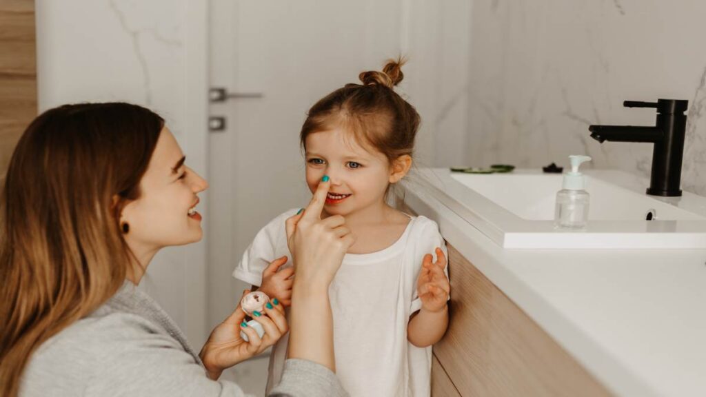 Een jonge vrouw met groene nagellak tikt speels op de neus van een jong meisje in een Scandinavische badkamer.